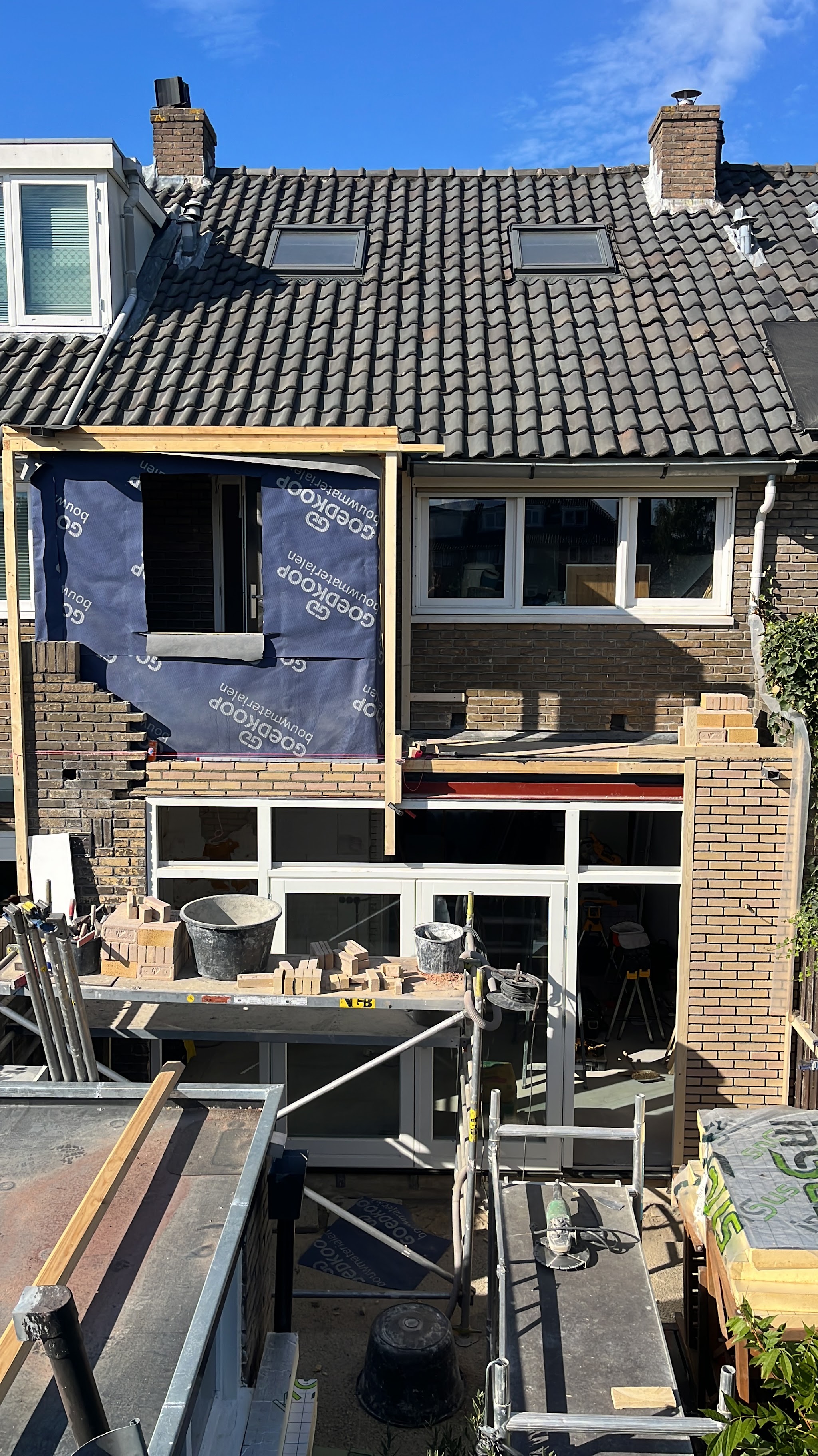 Achterzijde van een rijtjeshuis met een uitbouw en dakopbouw in aanbouw, steiger en stenen op de voorgrond